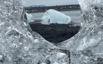 Blick durch ein rundes Loch in einem glasklaren Eisblock auf einen Eisberg am schwarzen Strand des Diamond Beach in Island.