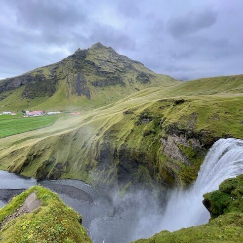 Blick von oben auf den mächtigen Skógafoss-Wasserfall in Island mit grünem Gras, steilen Hängen und Farmgebäuden im Hintergrund