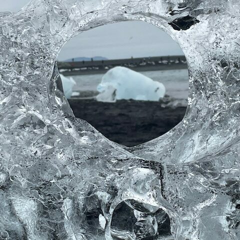 Blick durch ein rundes Loch in einem glasklaren Eisblock auf einen Eisberg am schwarzen Strand des Diamond Beach in Island.