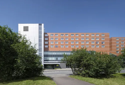 Hotelgebäude mit modernem Design, umgeben von Bäumen und blauem Himmel.