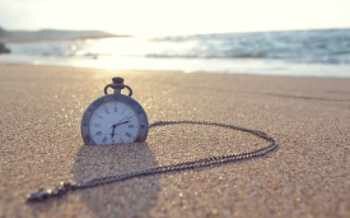 Eine Uhr, die im Sand gesteckt ist. Im Hintergrund sieht man das Meer.